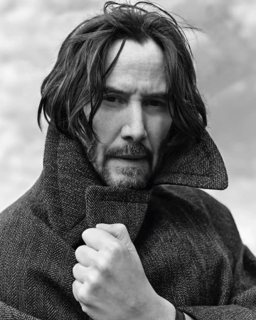 Black and white portrait of Keanu Reeves wearing a herringbone coat, looking at the camera with a neutral expression, against a blurred background.