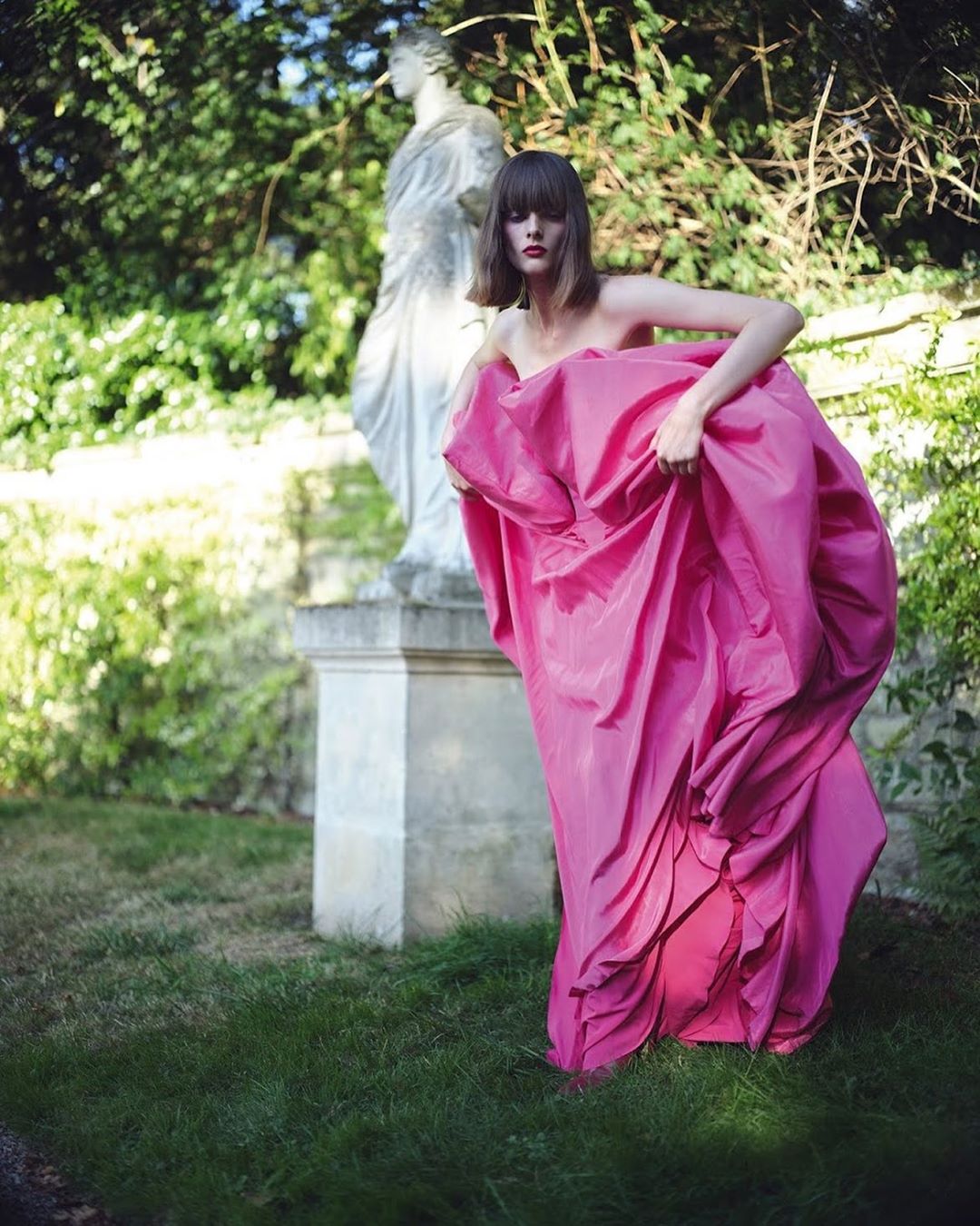 Woman in billowing pink fabric poses near statue in green garden setting under soft lighting. Fashion editorial with outdoor backdrop.