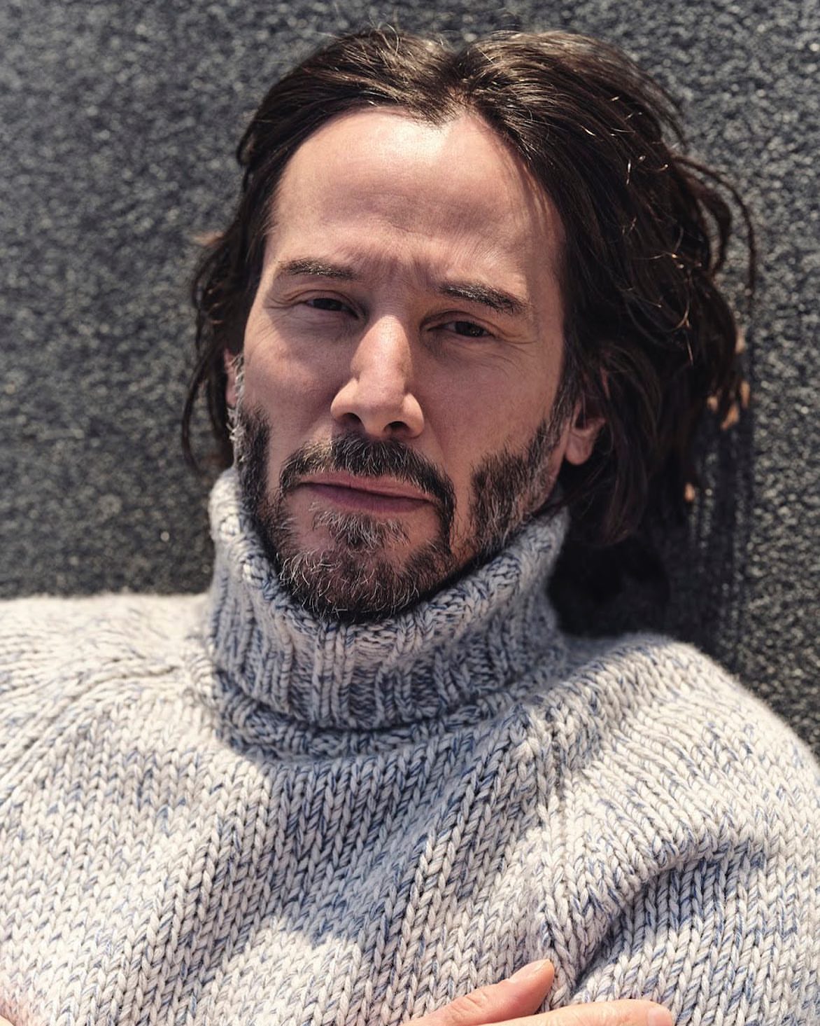 Close-up portrait of Keanu Reeves wearing a grey turtle neck sweater against a dark grey textured background, looking directly at the camera.