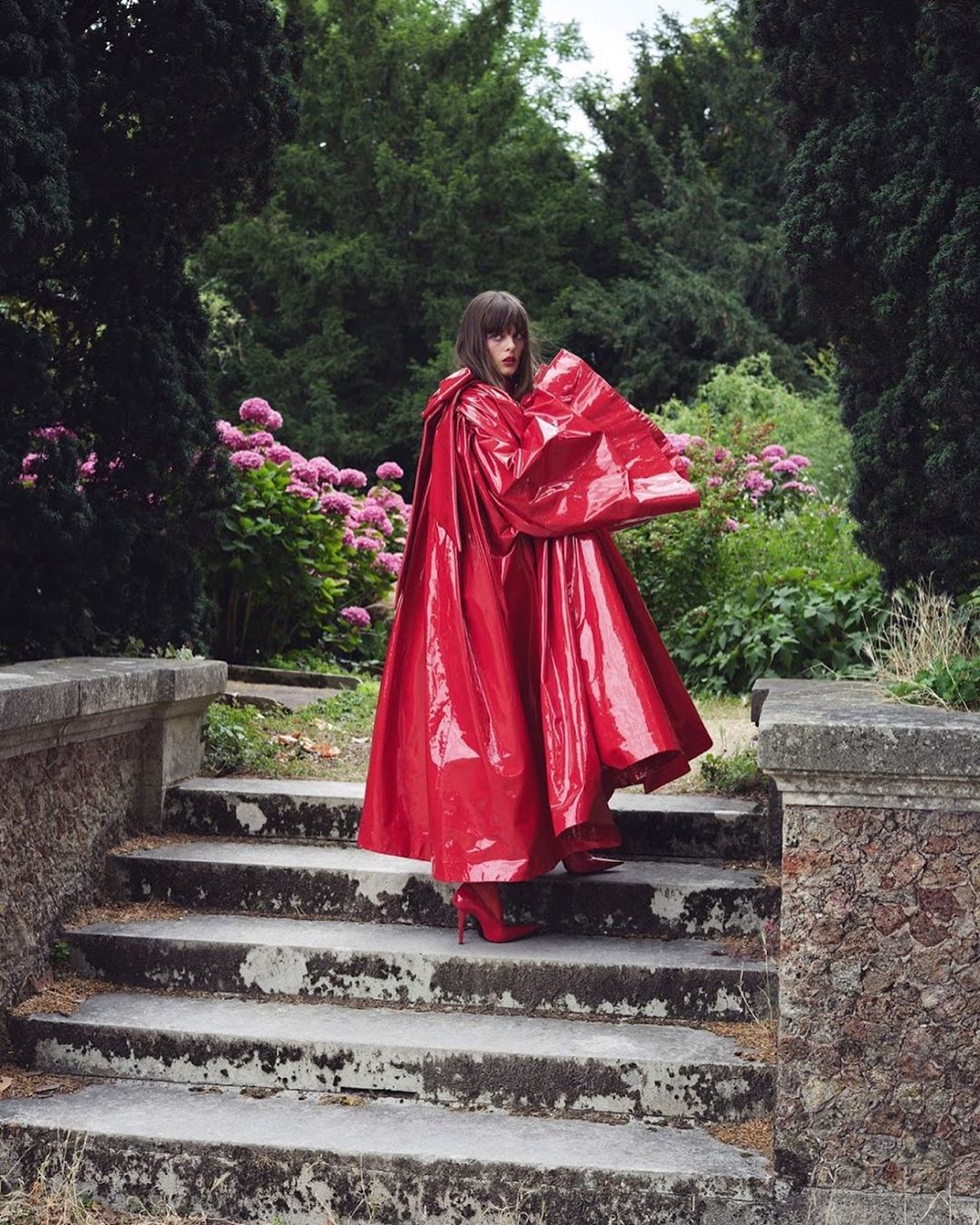 Woman in a red shiny cape and heels descending mossy stone steps in a garden with flowers and tall green trees in the background.