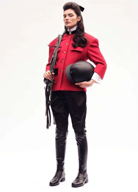 Full-length studio shot of a woman in a red riding jacket, black riding pants, and tall black boots holding a riding helmet.