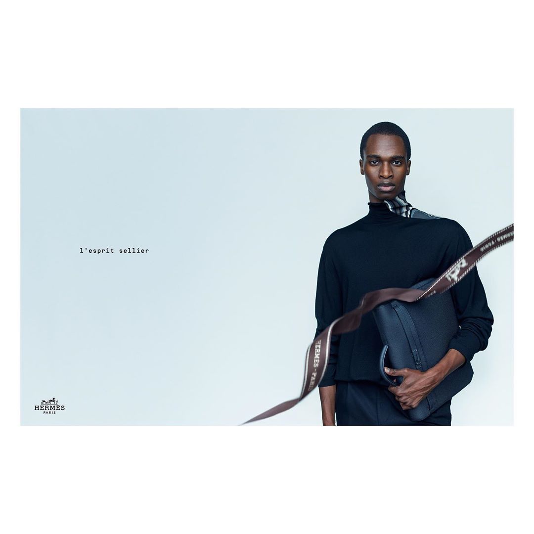 Black male model poses in a light studio with a modern black leather satchel and Hermes ribbon draped over his shoulder in a studio shot.