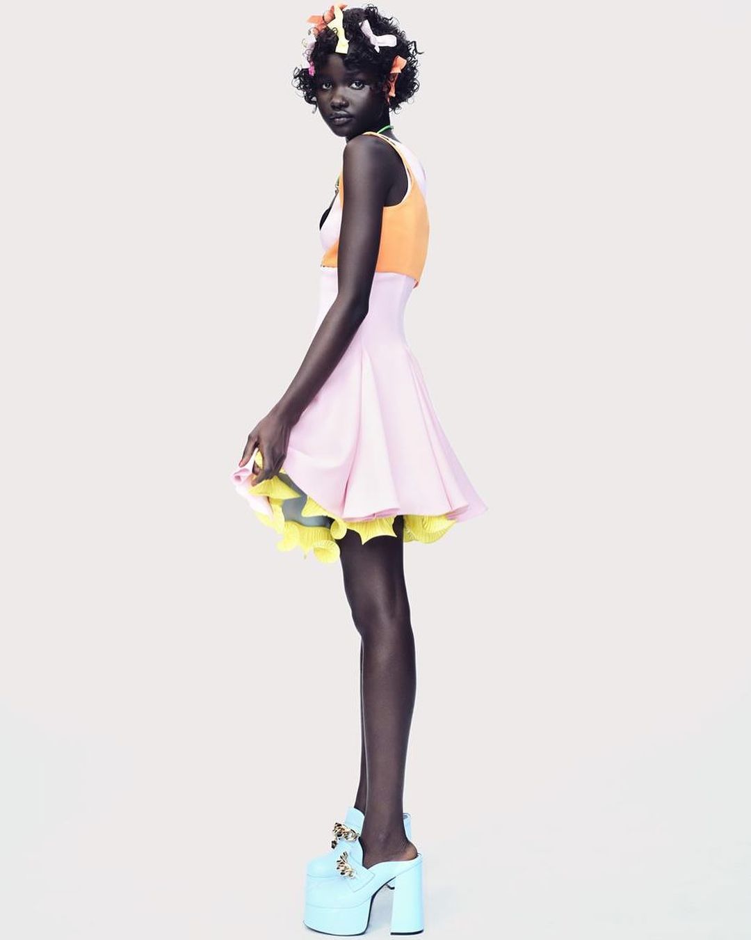 A slender African-American woman stands sideways in a pink and orange dress with large platform heels against a white backdrop in a studio shot.