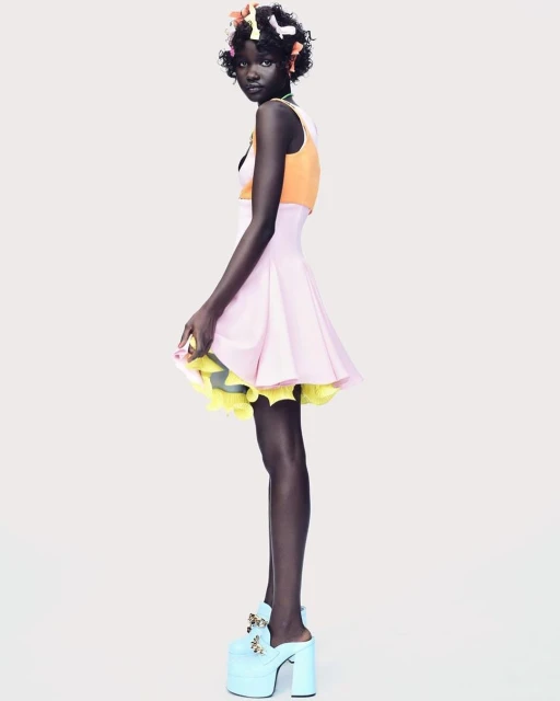A slender African-American woman stands sideways in a pink and orange dress with large platform heels against a white backdrop in a studio shot.