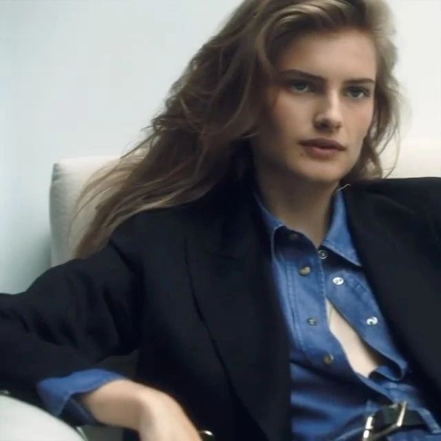 Elegant woman with long hair, wearing a denim shirt and black blazer, posing indoors in a fashion forward editorial shot.
