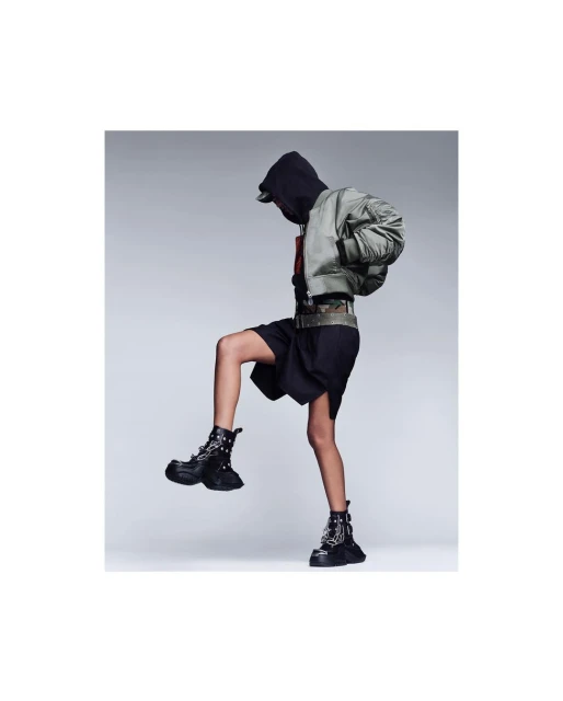 Person in a gray hooded jacket and black shorts wearing studded combat boots, one leg raised, in a neutral grey studio setting.