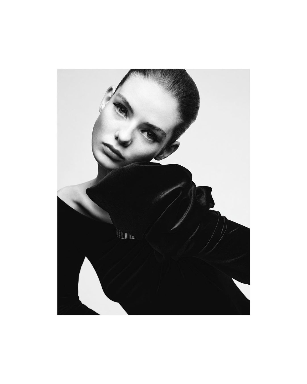 Monochrome portrait of a woman with slicked-back hair wearing a black dress with a large velvet bow, gazing at the camera in a studio setting.