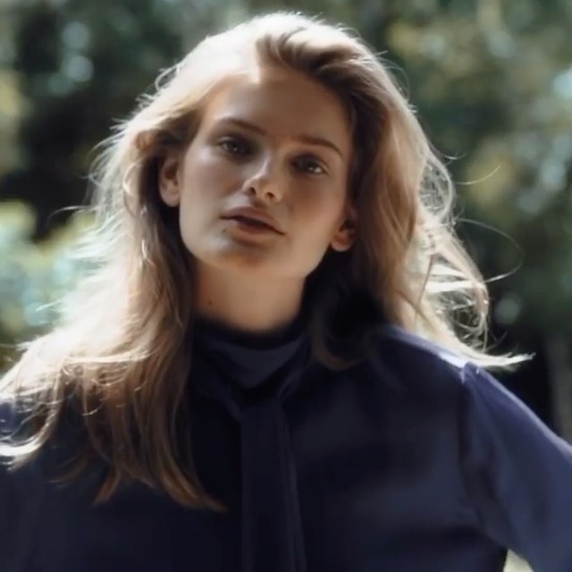 Portrait of a young woman with long blonde hair wearing a dark blue blouse outdoors with a softly blurred green background.