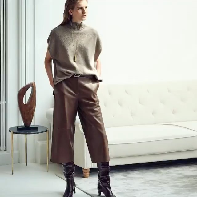 Indoor shot of a woman in a sleeveless top and leather pants, posing next to a small black table with an art piece on it.