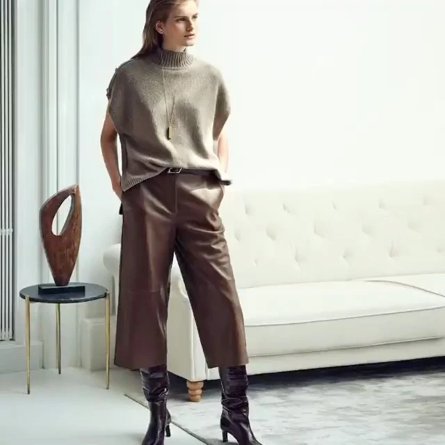 Indoor shot of a woman in a sleeveless top and leather pants, posing next to a small black table with an art piece on it.