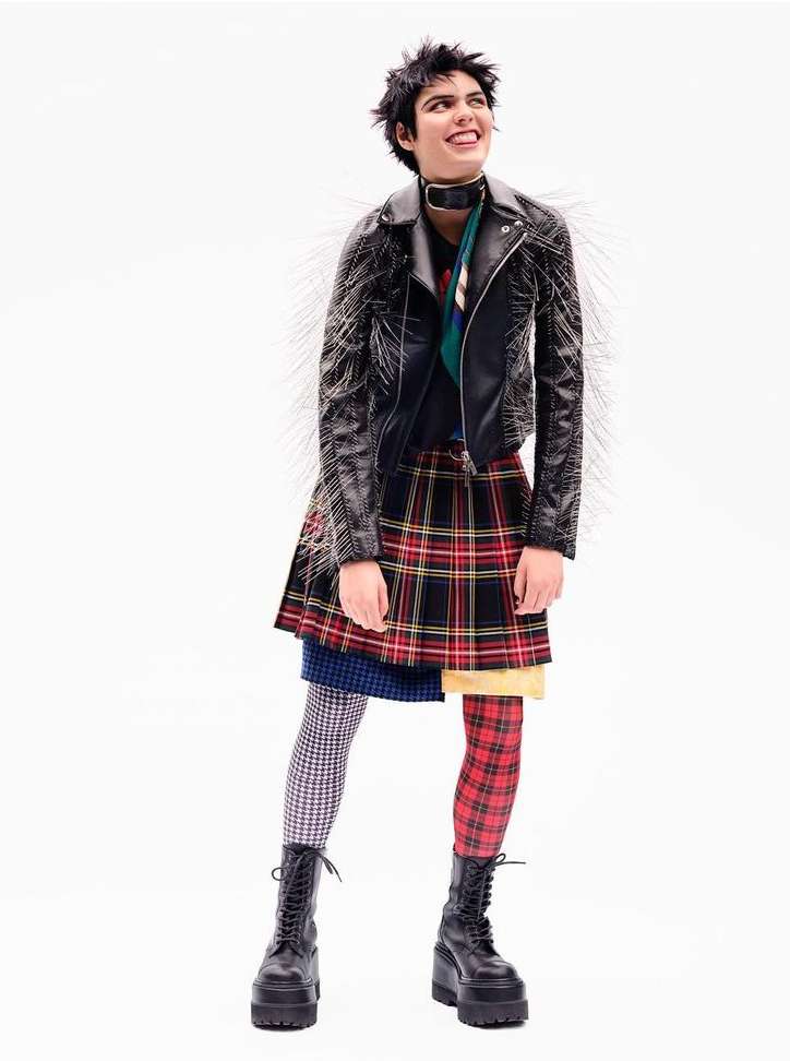 Full shot of a smiling woman with short dark hair wearing a studded black leather jacket, layered skirts, mismatched tights, and platform boots.