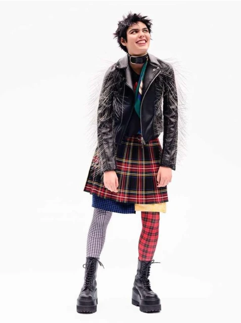 Full shot of a smiling woman with short dark hair wearing a studded black leather jacket, layered skirts, mismatched tights, and platform boots.