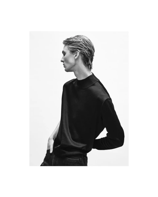 Monochrome portrait of a young adult male model posing in a dark sweater and jeans against a light background in a studio setting.