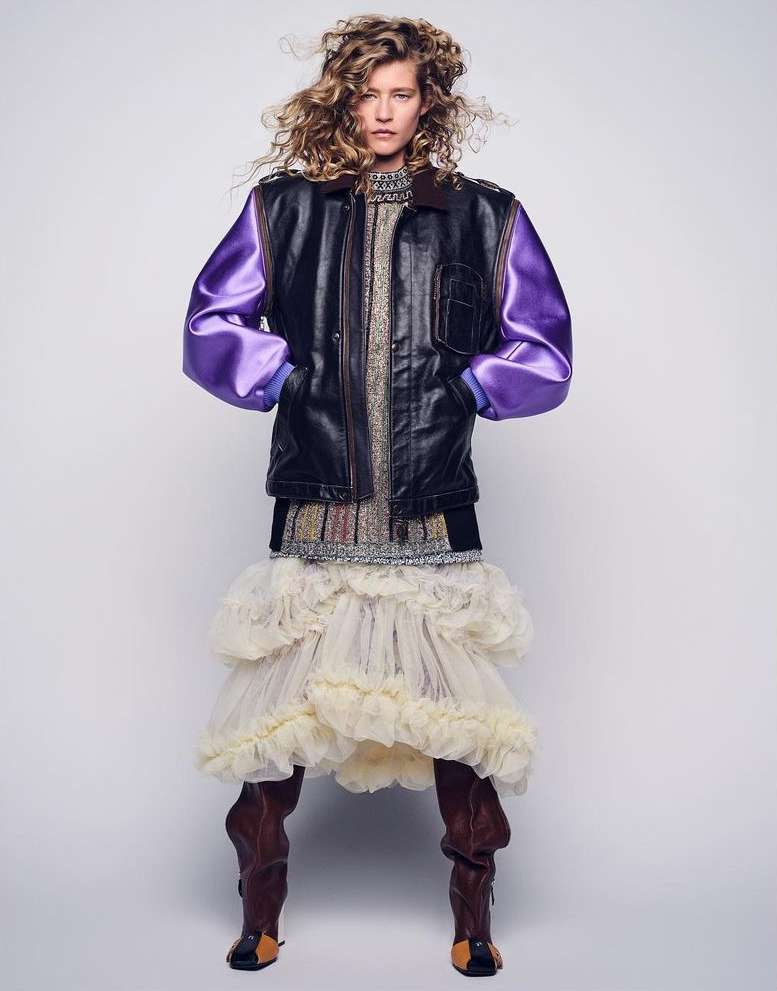 Model with curly blonde hair wearing a black and purple leather bomber jacket and ruffled cream skirt against a seamless light background