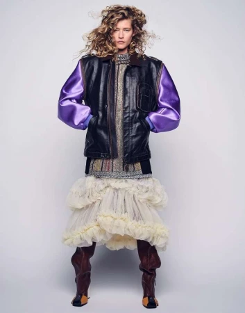 Model with curly blonde hair wearing a black and purple leather bomber jacket and ruffled cream skirt against a seamless light background