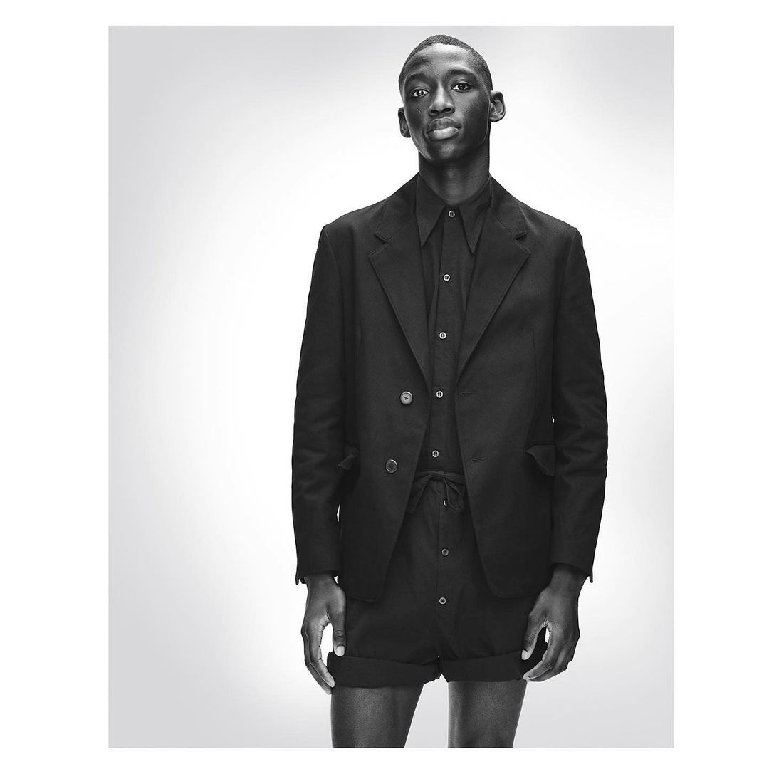 Monochromatic portrait of a Black male fashion model in a blazer, collared shirt, and tailored shorts against a gradient grey backdrop.
