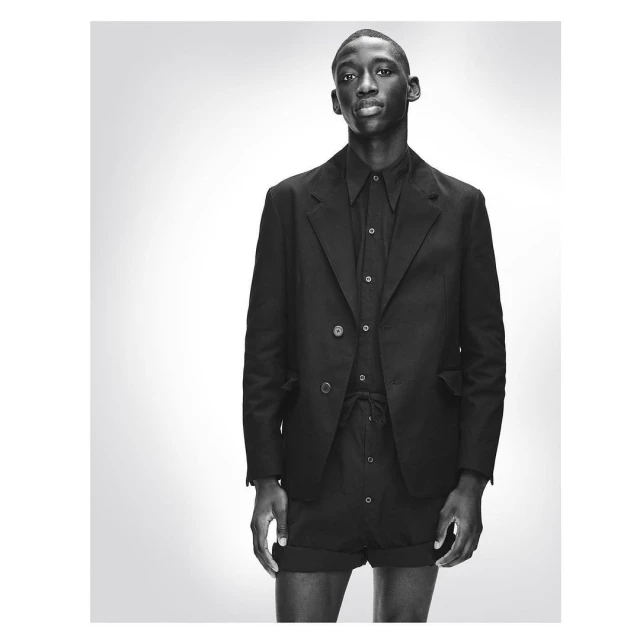 Monochromatic portrait of a Black male fashion model in a blazer, collared shirt, and tailored shorts against a gradient grey backdrop.