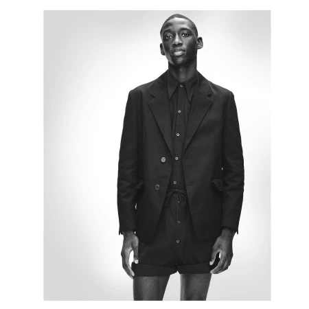 Monochromatic portrait of a Black male fashion model in a blazer, collared shirt, and tailored shorts against a gradient grey backdrop.