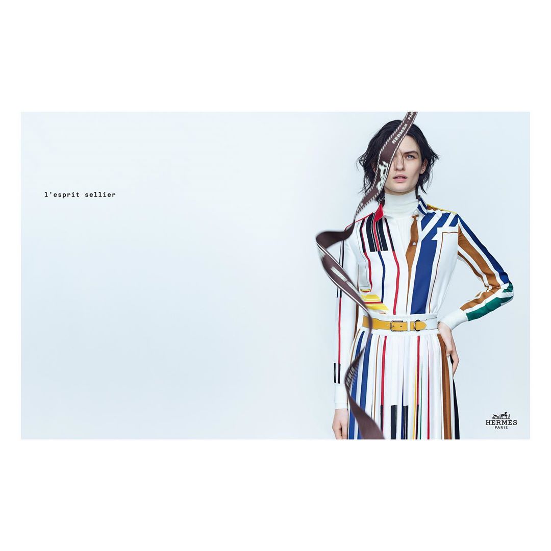 A woman in a colorful, striped dress with a yellow belt stands in front of a white background, partially obscured by a floating brown strap.