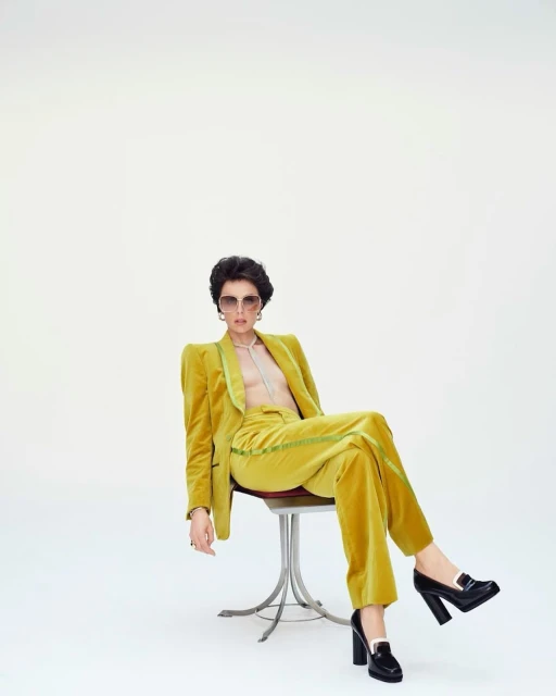 Woman in a yellow velvet suit sitting on a stool against a white backdrop, wearing sunglasses and black platform shoes.