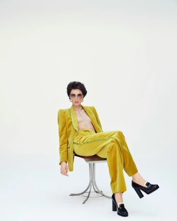 Woman in a yellow velvet suit sitting on a stool against a white backdrop, wearing sunglasses and black platform shoes.