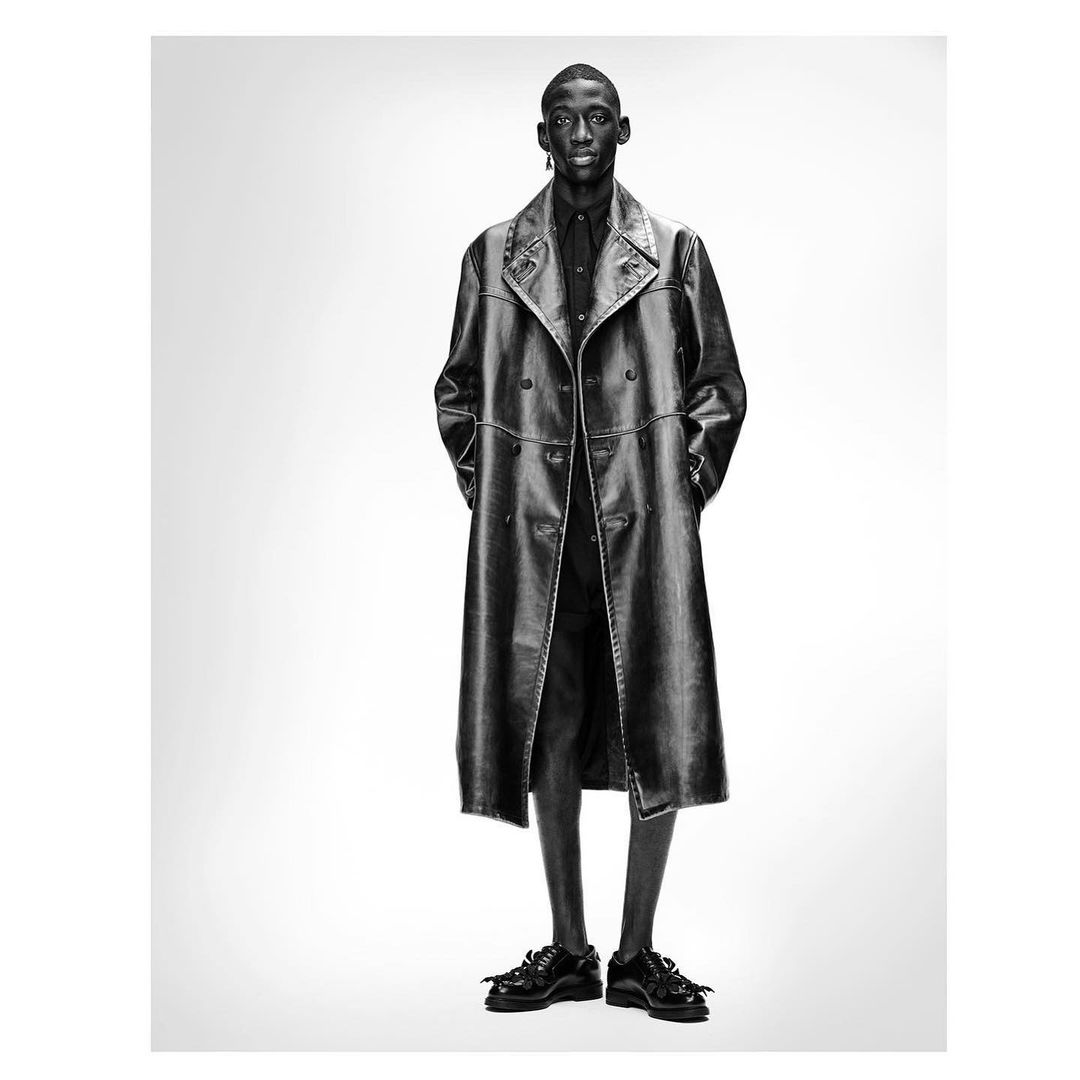 Full-length shot of a Black male model wearing a black leather trench coat, shirt, shorts, and shoes against a white backdrop in a studio setting.