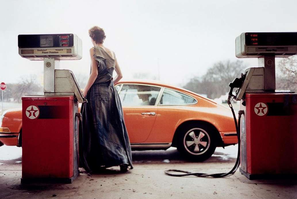 An adult female wearing a denim dress stands at a vintage Texaco gas pump with an orange classic Porsche 911 parked nearby outdoors.