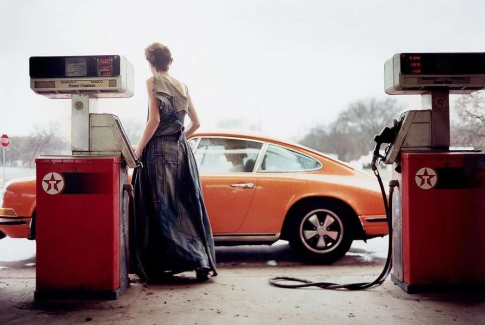An adult female wearing a denim dress stands at a vintage Texaco gas pump with an orange classic Porsche 911 parked nearby outdoors.