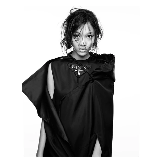 Black-and-white portrait of a young Asian woman in a black cape with a Prada logo, against a white background in a fashion editorial style.