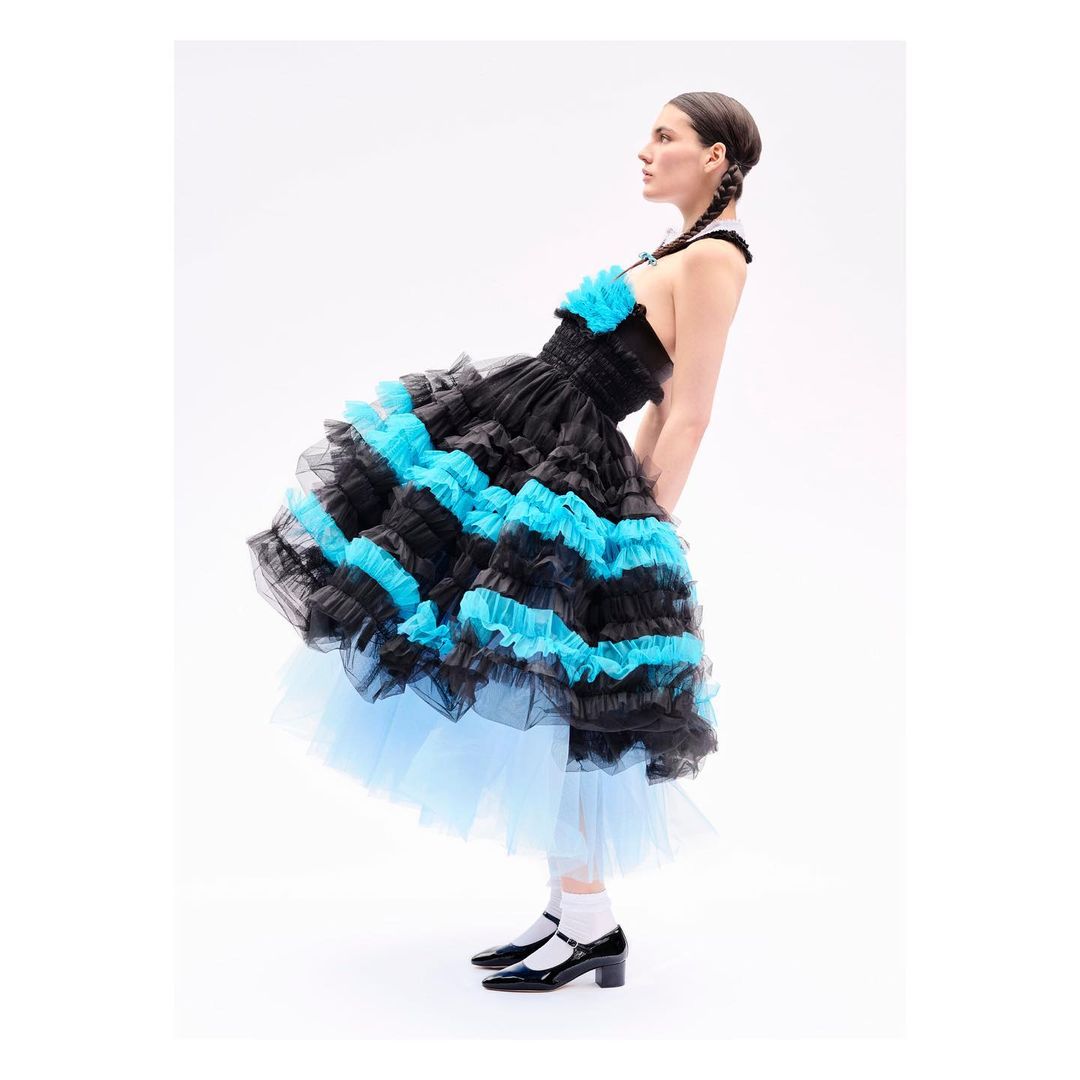 A young woman in a ruffled dress poses in a studio, with a white backdrop. The dress is primarily black with blue ruffles.