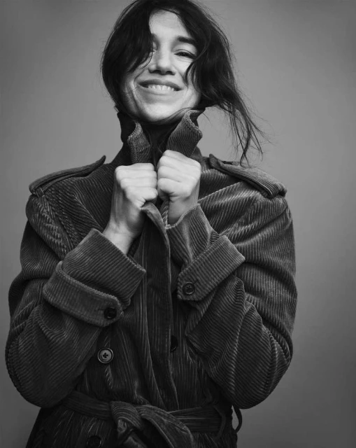 Black and white portrait of a smiling woman with dark hair wearing a corduroy trench coat, standing against a grey background.
