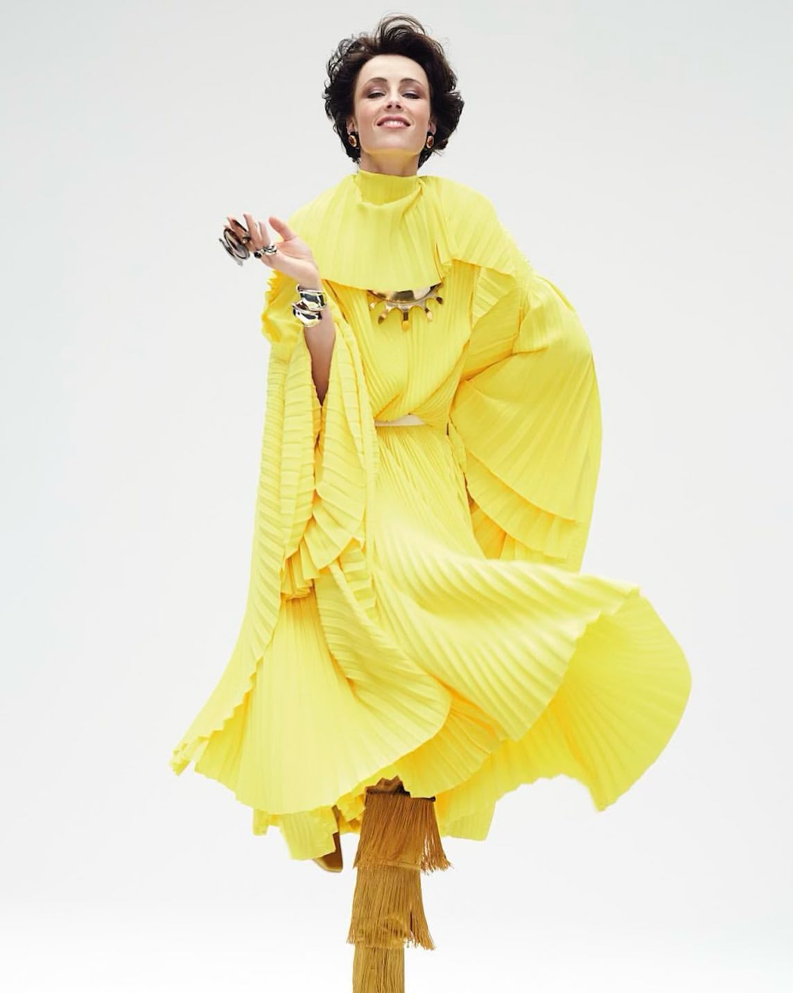 Woman wearing a yellow pleated dress with layered ruffles poses against a white background in a fashion editorial style studio shot.