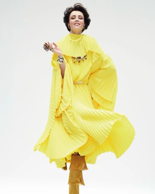 Woman wearing a yellow pleated dress with layered ruffles poses against a white background in a fashion editorial style studio shot.