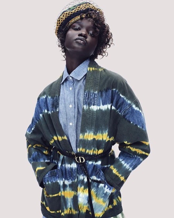 Fashionable Black woman poses in a tie-dye kimono-style jacket, blue collared shirt, and patterned headwrap, against a plain backdrop.