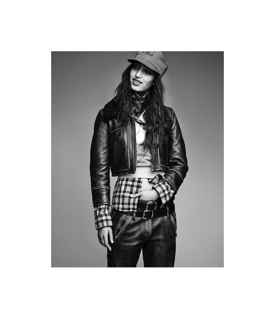 Monochrome portrait of a female model wearing a leather jacket, plaid shirt, and cap against a grey background.