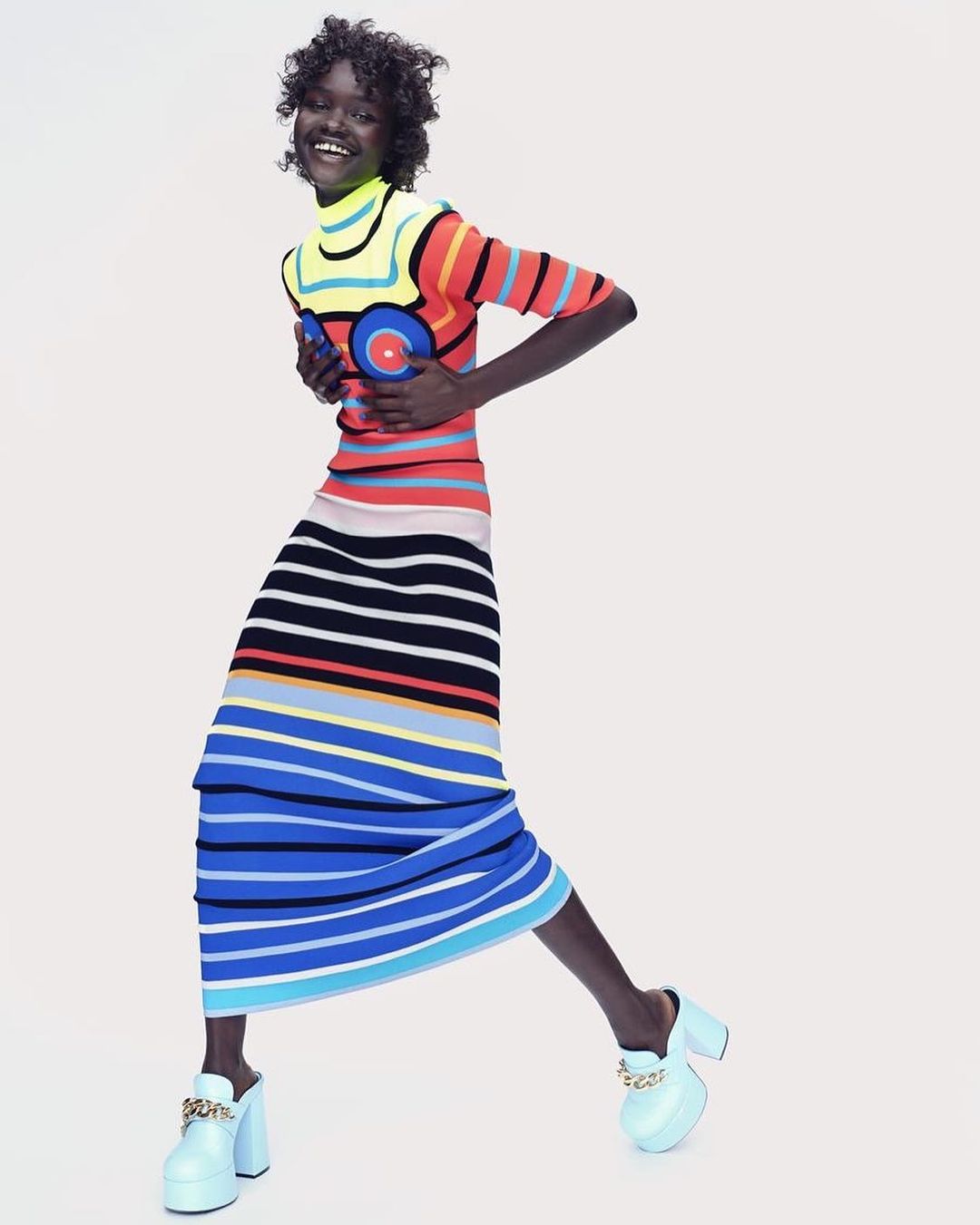 Woman smiles wearing a colorful striped dress and light blue platform heels against a plain background in a fashion studio shot.