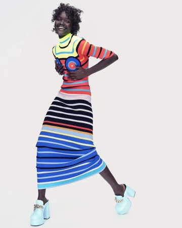 Woman smiles wearing a colorful striped dress and light blue platform heels against a plain background in a fashion studio shot.