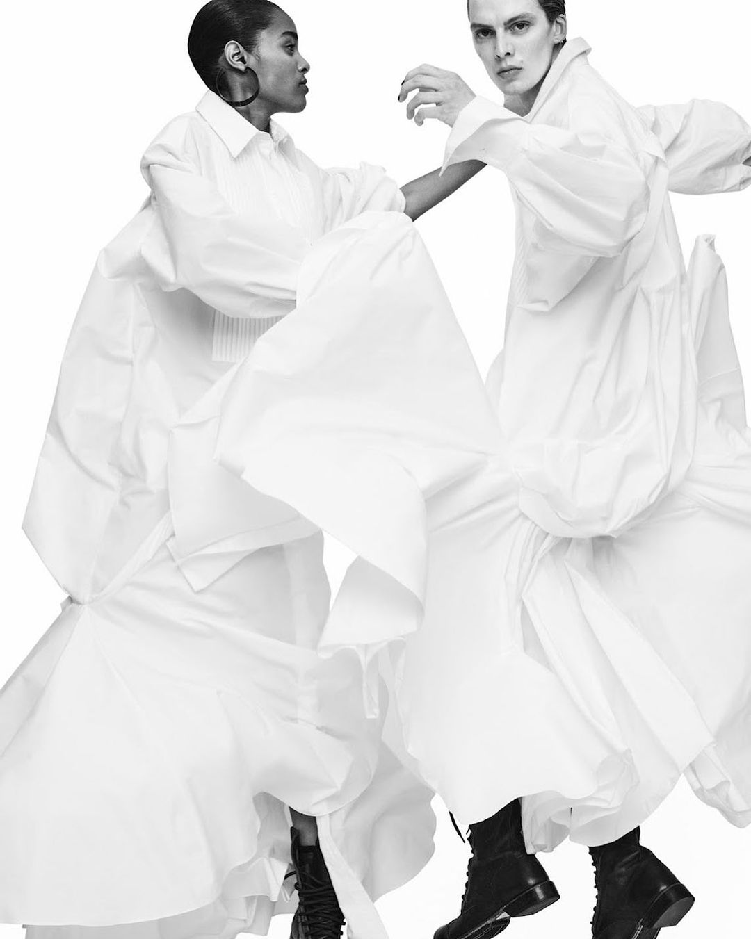 Two fashion models, one Black and one White, pose in flowing white dresses and black boots against a stark white background in studio lighting.