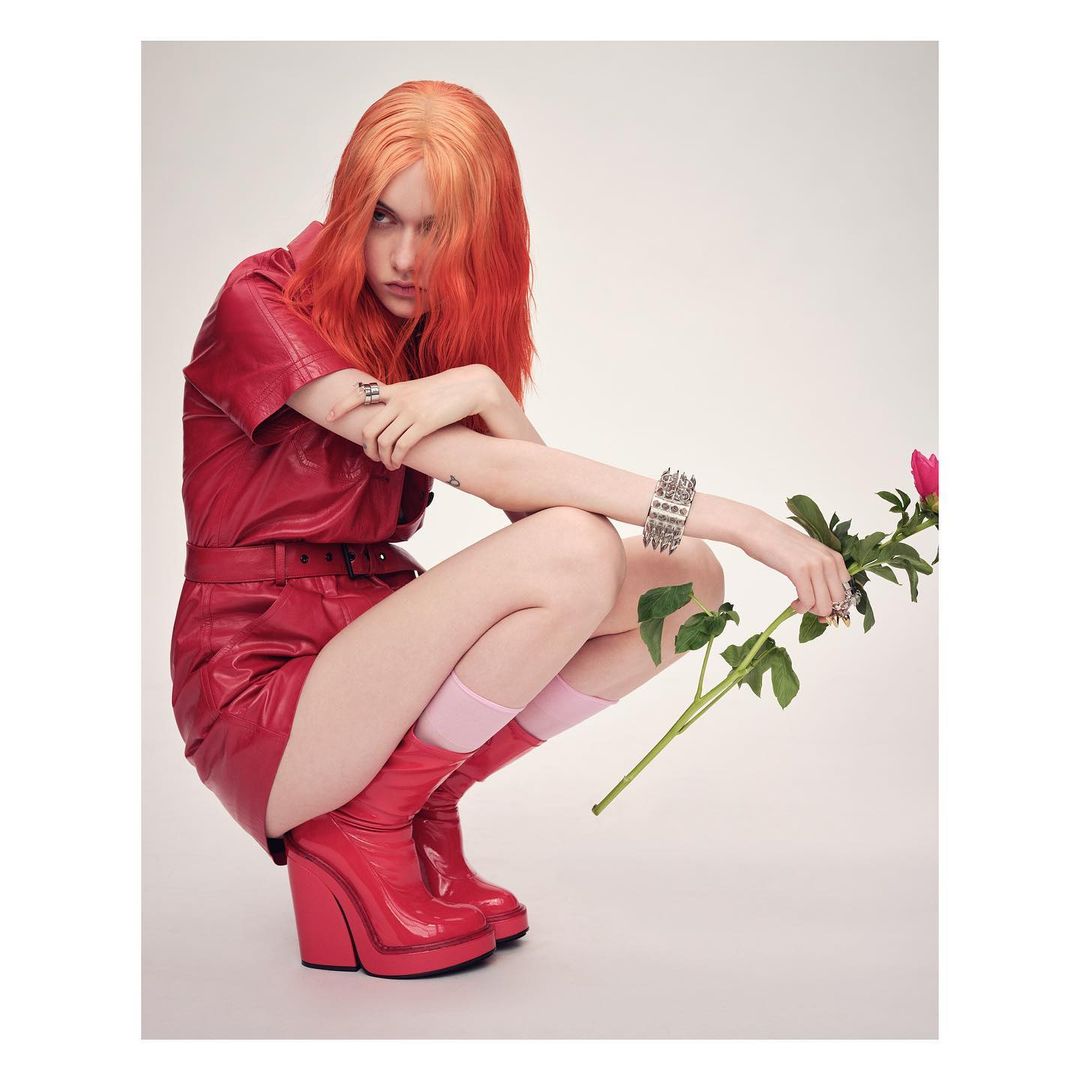 Young woman with red hair in red leather romper and boots, kneeling indoors holding a pink peony, against a plain white background.