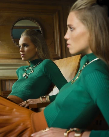Adult female portrait looking at reflection in mirror. She wears a green turtleneck top, jewelry, and a gold skirt, indoors.