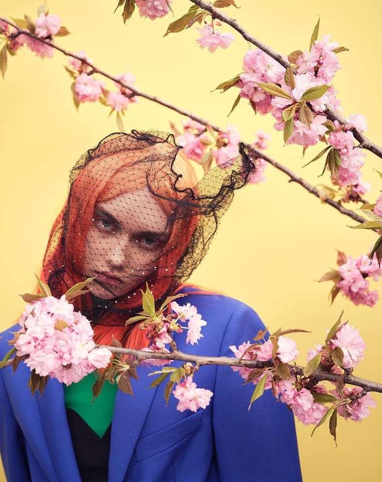 A woman with orange hair and a black veil poses near blooming pink cherry blossoms against a bright yellow backdrop in a fashion shot.