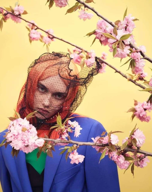 A woman with orange hair and a black veil poses near blooming pink cherry blossoms against a bright yellow backdrop in a fashion shot.