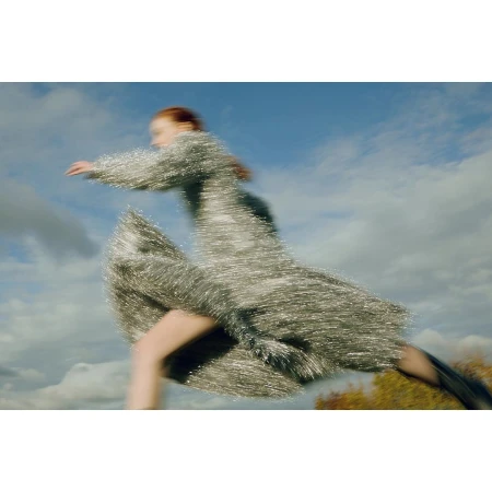 A motion-blurred female model wearing a metallic fringed dress running outdoors against a cloudy blue sky.