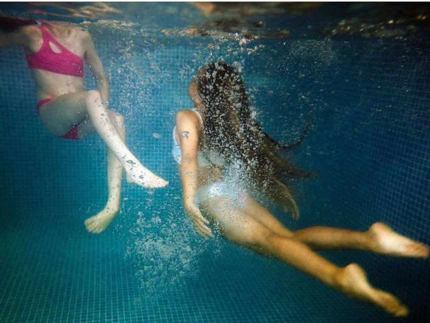Two young women underwater in a pool with blue tile, sunlight filtering through water, creating a refreshing and dynamic scene.