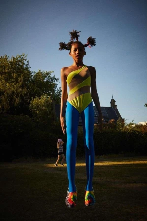 Young girl in a yellow and blue swimsuit posing in a field against a blue sky, trees, and a house with solar panels in the background.