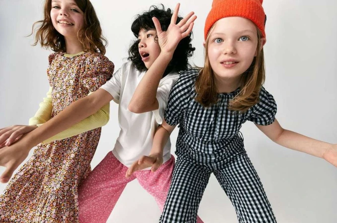 Three smiling girls in fashionable outfits strike a dynamic pose against a white backdrop, showcasing a playful fashion-forward style.