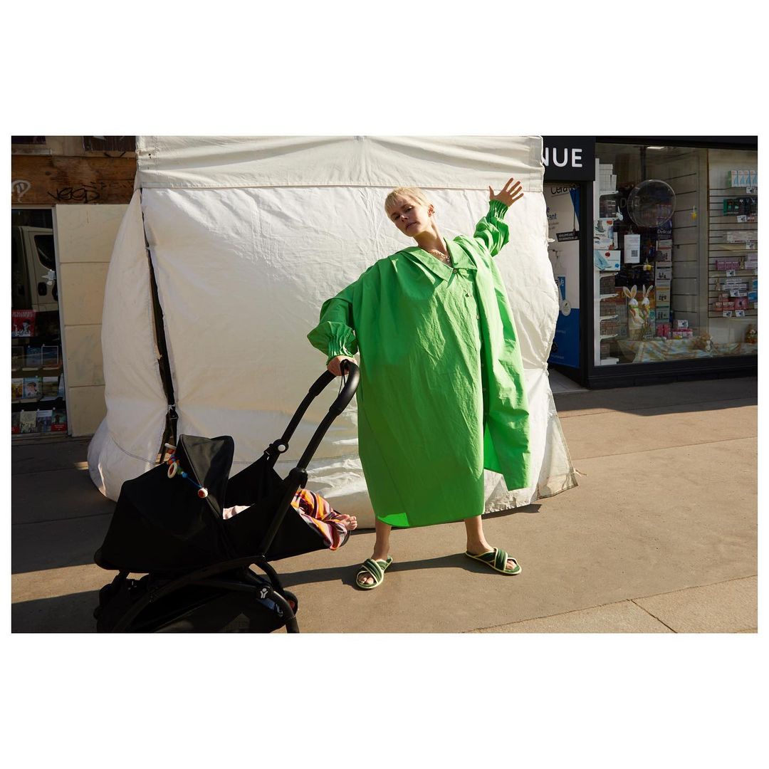 Adult female in an oversized green dress pushes a baby stroller on a sidewalk in front of a store. The woman waves with her right arm.