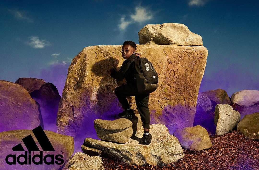Young boy climbs a rock formation wearing an Adidas backpack and athletic wear, set against a blue sky with the Adidas logo visible.
