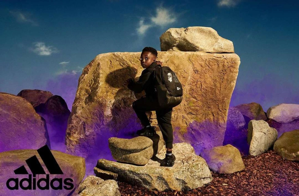 Young boy climbs a rock formation wearing an Adidas backpack and athletic wear, set against a blue sky with the Adidas logo visible.