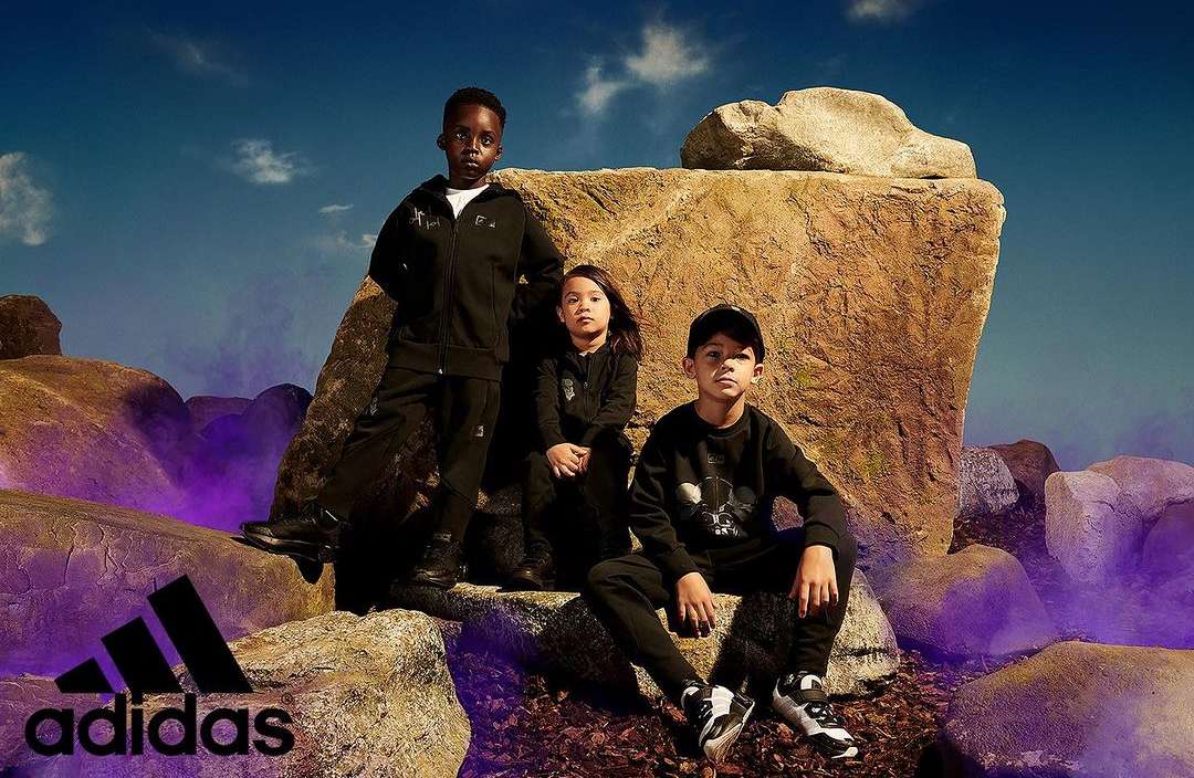 Three children in black Adidas tracksuits pose on rocks against a blue sky, showcasing sportswear and outdoor fashion in a vibrant setting.